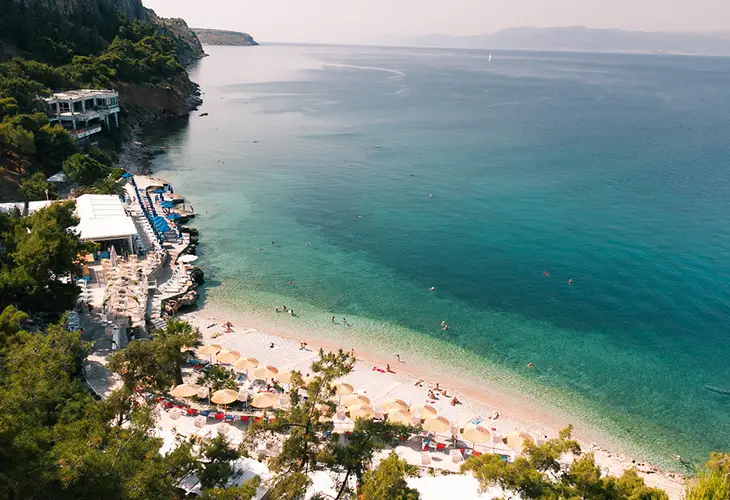 A rocky beach, with a beach bar right on top, clean waters and a view of the Argolic Gulf