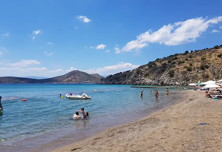 A beach with a mixture of pebble and sand, with families playing in the water and umbrellas in the background