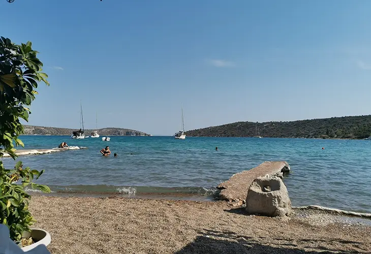 A small pebble cove with a short concrete jetty, and two sailing boats deeper in the waters